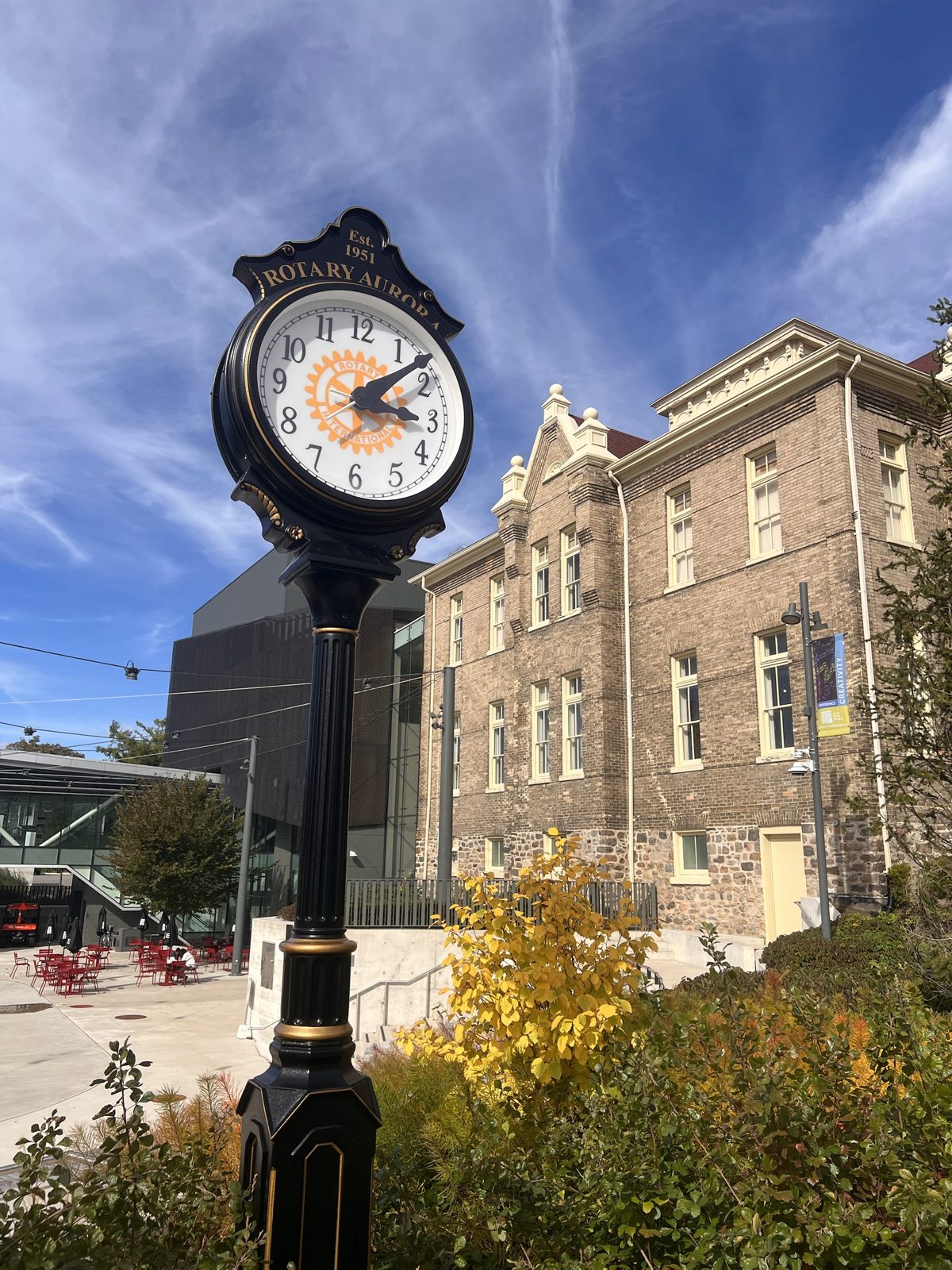 The black-and-gold Rotary Aurora street clock, established 1951, beside the historic brick Church Street School under a blue autumn sky